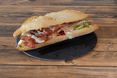 Domuz pastırmalı sandviç ve siyah tabakta marul. Sandviç ikiye bölünür ve ahşap bir masaya yerleştirilir.