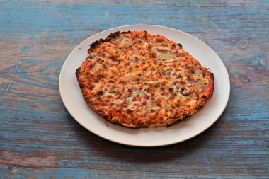 Bir pizza ahşap bir masada beyaz bir tabağın üzerinde oturuyor. Pizza peynirle kaplı ve birkaç malzemesi var. Tabak temiz ve pizza lezzetli görünüyor.