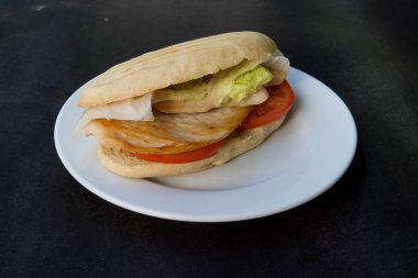 Beyaz tabakta salatalı ve domatesli bir sandviç. Sandviç ikiye bölünür ve siyah bir masaya yerleştirilir.