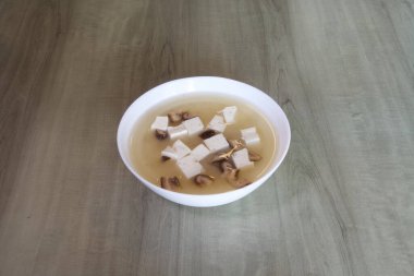 Tofu ve mantarlı bir kase çorba. Çorba beyaz ve kremalı bir dokusu var. Kase ahşap bir masaya konur.