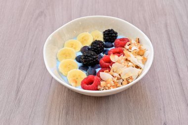 Bir kase yaban mersinli, muzlu ve ahududulu meyve. Kase beyaz ve tahta bir masaya yerleştirilmiş.