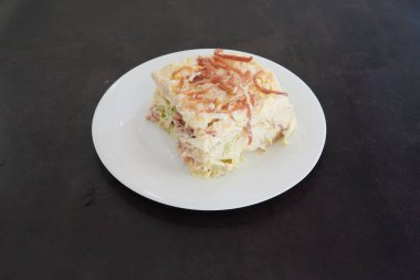 Bir dilim yiyecek beyaz bir tabakta duruyor. Yemek, pastırma ve peynirli katmanlı bir yemektir.