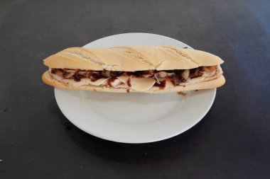 Beyaz tabakta bir sandviç var. Sandviç et ve peynirden yapılmıştır. Tabak masanın üstünde.