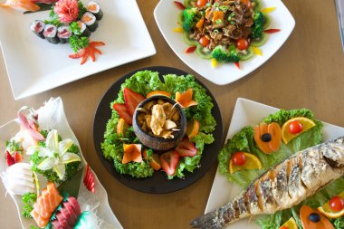 Suşi, balık ve sebzeler de dahil olmak üzere çeşitli Asya yemekleri bir masada düzenlenir. Sunum renkli ve iştah açıcı, her yemek taze ve lezzetli görünüyor.