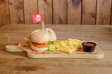 Üstünde kırmızı bayrak ve yanında patates kızartması olan bir hamburger. Hamburger ahşap bir tahtanın üzerinde, altında kırmızı beyaz bir kağıt var.
