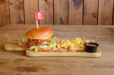 Üstünde kırmızı bayrak ve yanında patates kızartması olan bir hamburger. Hamburger ahşap bir tahtanın üzerinde ve patates kızartması da siyah bir kabın içinde.