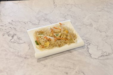 Sebzeli bir tabak erişte. Tabak beyaz ve sebzeler turuncu. Tabak mermer bir tezgahın üzerinde.