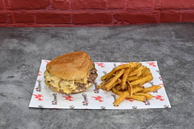 Kırmızı beyaz kağıtta hamburger ve patates kızartması. Hamburger peynirle kaplıdır ve iki köftesi vardır. Kızartmalar baharatlı ve çıtır çıtır görünüyor.