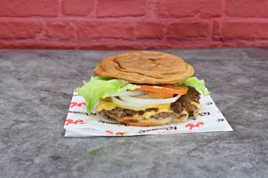 Kırmızı peçeteye salatalı, domatesli ve soğanlı bir hamburger. Hamburger, arka planda tuğla duvar olan bir masada.