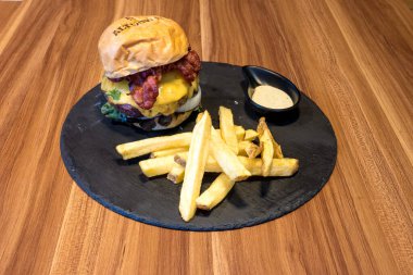 Siyah tabakta hamburger ve patates kızartması. Hamburgerin üstünde peynir ve pastırma var. Kızartmalar altın sarısı ve çıtır çıtır. Tabak ahşap bir masaya konmuş.