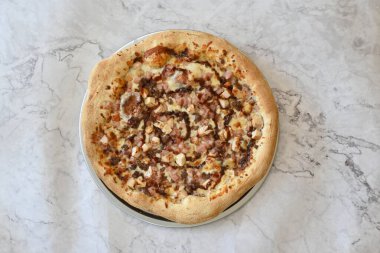 Jambonlu ve tavuklu bir pizza mermer tezgahın üzerinde duruyor. Pizza peynirli ve pastırmalı ve lezzetli görünüyor.