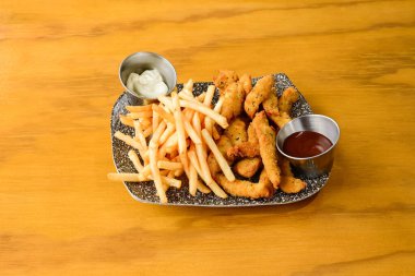 Tahta bir masada bir tabak tavuk kanadı ve patates kızartması duruyor. Kızartmalar altın ve çıtır çıtır, tavuklar pişti ve yenmeye hazır. Küçük bir kap ketçap.