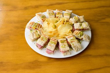 Tahta bir masanın üzerinde bir tabak sandviç ve cips var. Sandviçler küçük parçalara ayrılır ve tabakta daire şeklinde dizilir. Çipler sandviçlerin etrafına saçılmış.