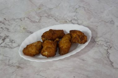 Bir tabak kızarmış tavuk kanadı mermer tezgahın üzerinde duruyor. Kanatları altın sarısı ve çıtır çıtır görünüyor. Tabak beyaz ve tezgah mermer, sahneyi temiz gösteriyor.