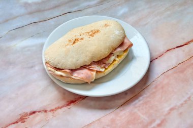 Beyaz tabakta jambonlu sandviç var. Sandviç ikiye bölündü. Tabak mermer tezgahın üzerinde.