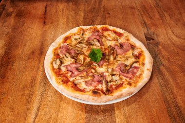 Mantarlı bir pizza ve ahşap bir masanın üstünde jambon. Pizza dilimlenir ve beyaz bir tabakta servis edilir.