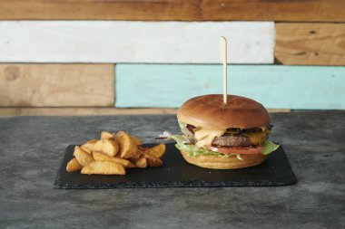 İçinde kürdan olan bir hamburger siyah bir tabakta duruyor. Hamburger peynirli ve bir dilim domatesli. Bir de patates kızartması var.