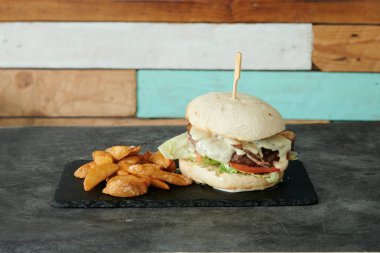 Üzerinde marul ve domates olan bir hamburger siyah bir tabakta duruyor. Tabakta patates kızartması da var.
