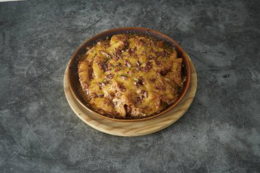 Peynirli ve etli bir kase yemek. Kase ahşap bir tabakta. Tabak gri bir yüzeyde.