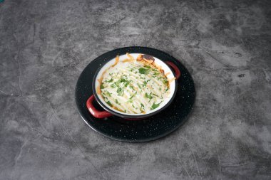Beyaz soslu ve yeşil otlu lezzetli bir yemek siyah bir tabağın üzerinde duruyor. Çanak gri bir yüzeye yerleştirilmiş. Tezgah ya da masa olabilir.