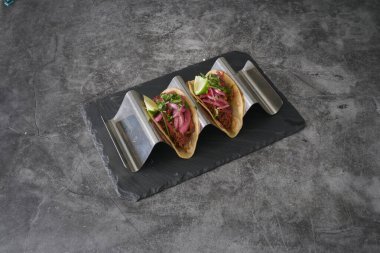 Siyah tabakta limonlu ve soğanlı iki taco. Tabak gri bir yüzeyde. Tacolar metal bir tepside.
