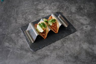 Siyah tabağın üstünde limonlu iki taco. Tabak gri bir yüzeyde. Tacolar metal bir tepside.
