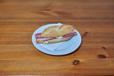 Beyaz tabakta bir sandviç var. Tabak ahşap bir masanın üzerinde. Sandviç ikiye bölündü.