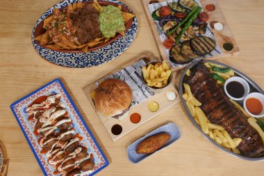 İçinde sandviç, patates kızartması ve biftek de olan çeşitli yiyecekler olan bir masa. Ayrıca bir kase sos da var.