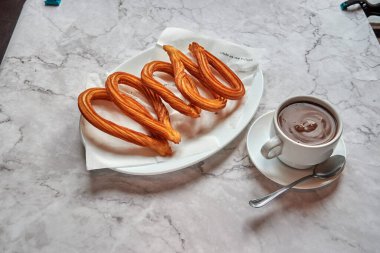 Bir tabak lezzetli churros bir peçetenin üzerinde oturuyor, yanında da bir fincan sıcak çikolata var. Churro 'lar altın sarısı kahverengidir ve taze yapılmış gibi görünürken sıcak çikolata buharlaşır ve davetkar olur.