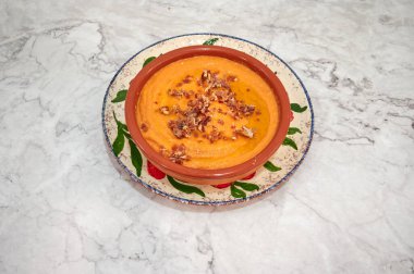 Üstünde pastırma parçaları olan bir kase çorba. Kase beyaz bir tabakta. Çorba turuncu renkli.