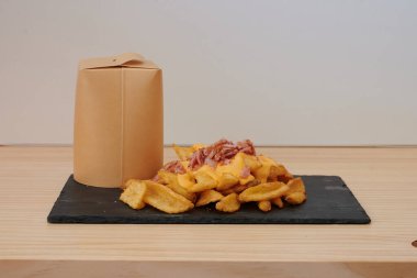 Bir kutu patates kızartması bir bardağın yanındaki masada oturur. Kızartmalar peynir ve pastırmayla kaplı.