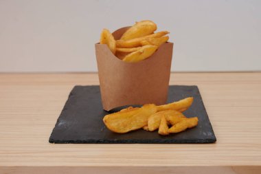 Tahta bir masada patates kızartması konteynırı duruyor. Patatesler ikiye bölünmüş ve kese kağıdına konmuş.