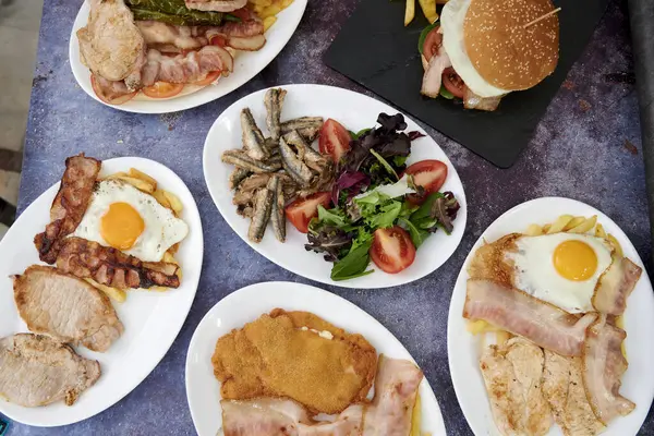 Pastırma, yumurta ve sandviç tabakları olan bir masa. Masada biraz sebze de var.