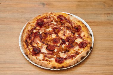 Sucuklu ve jambonlu bir pizza. Pizza beyaz bir tabakta. Tabak ahşap bir masanın üzerinde.