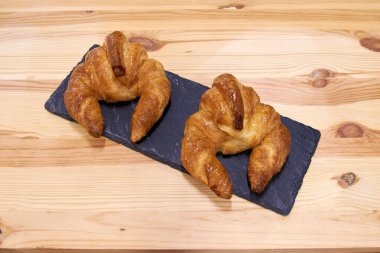 Siyah bir tabakta iki kruvasan. Tabak ahşap bir masanın üzerinde. Kruvasanlar altın kahverengidir ve tatlı bir jöleye sahiptir.