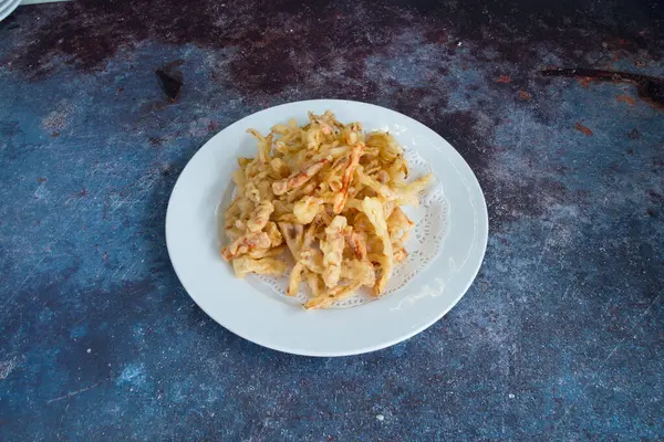 Mavi tezgahın üzerinde bir tabak kızarmış yiyecek var. Tabak beyaz ve altında peçete var.