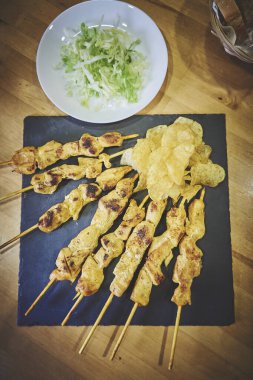 Yanında cips olan bir tabak tavuk şişi. Şişler siyah bir tabakta, cipsler de kasenin içinde.