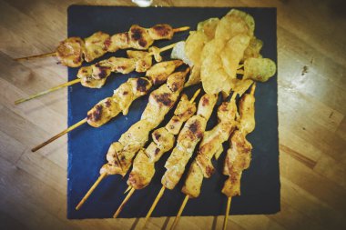 Yanında cips olan bir tabak tavuk şişi siyah bir tabağa konur. Şişler uzun ve incedir, bazılarının hafif kömürleşmiş bir görüntüsü vardır.