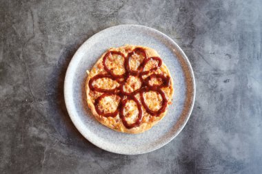 Üstünde barbekü sosu olan bir pizza. Sos bir çiçek şeklindedir. Pizza beyaz tabakta.