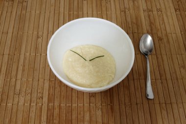 Kremsi beyaz bir madde ile dolu beyaz bir kase ahşap bir masada oturur. Kasenin kenarında kullanılmaya hazır gümüş bir kaşık duruyor. Kaşık kasenin arkasına yerleştirilmiş.