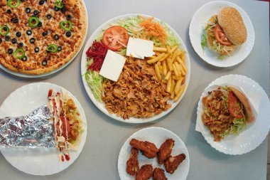 İçinde pizza, tavuk kanadı ve sandviç de olan bir masa. Masada birkaç tabak da yemek var.