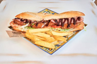 Beyaz tabakta pastırmalı ve domatesli bir sandviç. Patates kızartması. Sandviç ikiye bölündü.