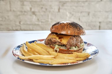 Peynirli ve pastırmalı bir hamburger patates kızartmasıyla birlikte bir tabakta oturur. Patatesler ince dilimlenmiş ve hamburgerin etrafına yerleştirilmiş.