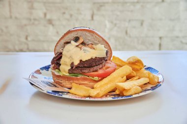 Ekmeğin üstünde peynir ve mantarlı bir hamburger. Patates kızartması. Hamburger beyaz tabakta.