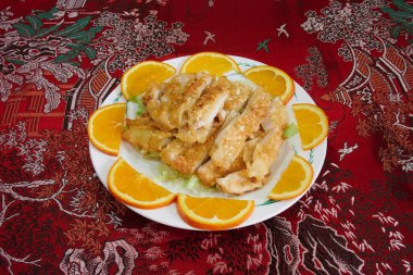 Üstünde portakal dilimleri olan bir tabak tavuk. Tabak kırmızı bir masa örtüsünün üzerinde. Tavuk parçalara ayrılır ve yanında portakal dilimleri ile servis edilir.