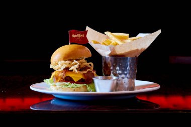 Soğanlı ve peynirli bir hamburger tabakta oturur. Tabakta küçük bir kap ketçap var.