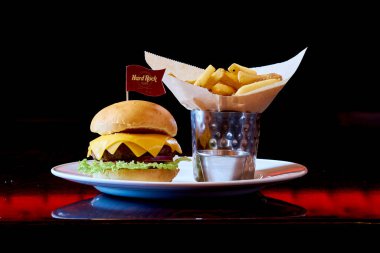 Beyaz tabakta peynirli ve marullu bir hamburger. Hamburgerin üstünde bayrak var. Hamburgerin yanında bir kase patates var.