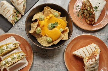 Bir masa burrito, taco ve nachos gibi çeşitli Meksika yemekleri ile doludur. Bir kase nachos ve guacamole de dahil olmak üzere farklı yemek türleri olan birkaç kase ve tabak var.