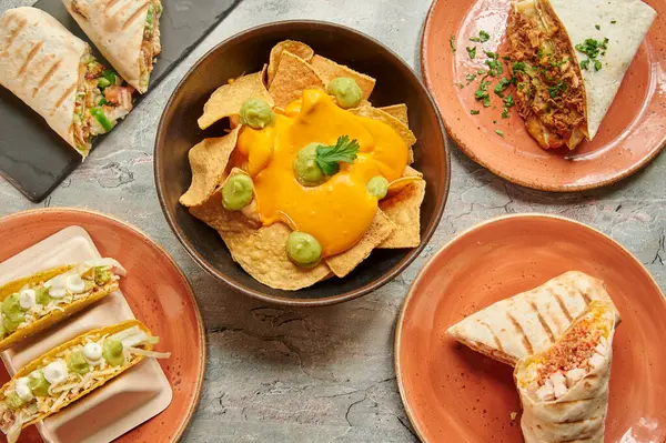 Bir masa burrito, taco ve nachos gibi çeşitli Meksika yemekleri ile doludur. Bir kase nachos ve guacamole de dahil olmak üzere farklı yemek türleri olan birkaç kase ve tabak var.