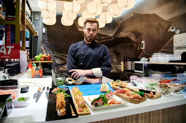 Şef üniformalı bir adam tezgahın arkasında çeşitli suşi ve diğer Asya yemekleriyle duruyor.